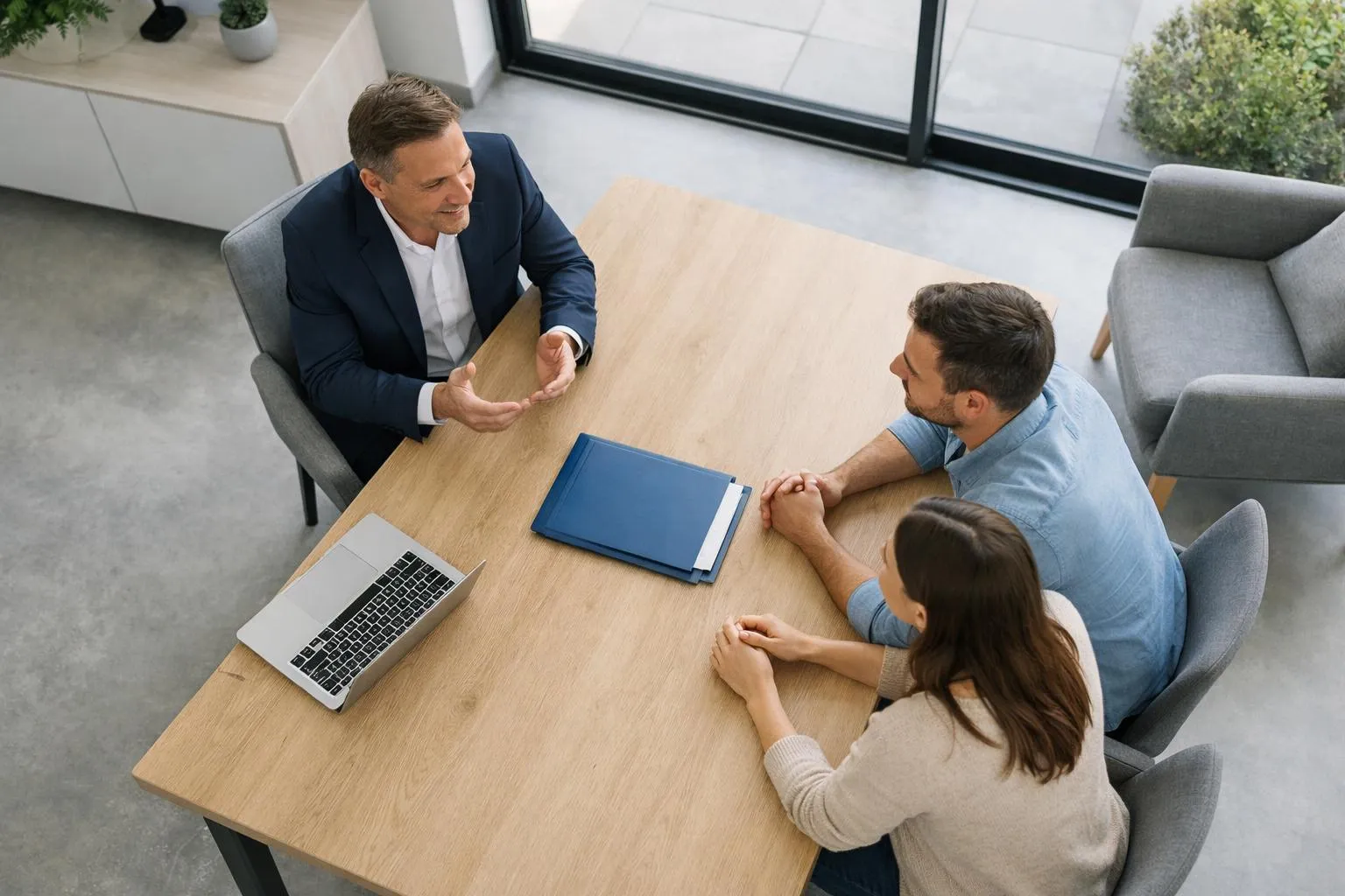 Banquier français assis face à un couple trentenaire dans un bureau moderne, posant un dossier confidentiel sur le bureau en bois clair, expression sérieuse et professionnelle, lumière naturelle par grande fenêtre, décor élégant avec plante verte, ambiance de négociation immobilière importante, aucun texte visible